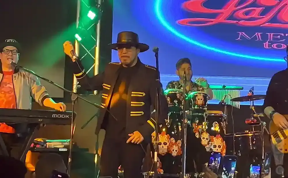 A band performs on stage, with a singer in a black hat and jacket holding a microphone, surrounded by other musicians.