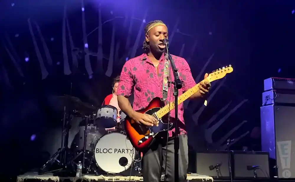 A Black man in a pink patterned shirt plays a sunburst electric guitar on stage with a drummer in the background.