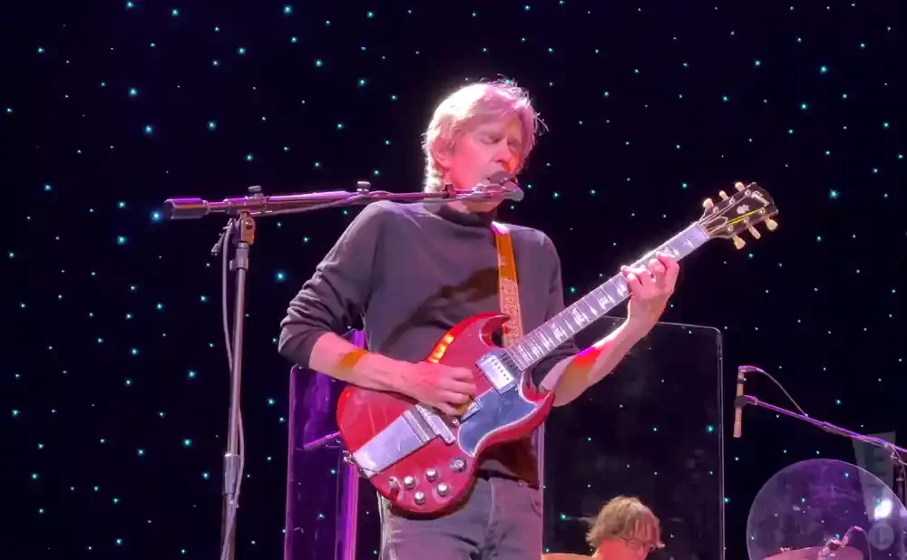 A man plays a red guitar on stage with a starry background.