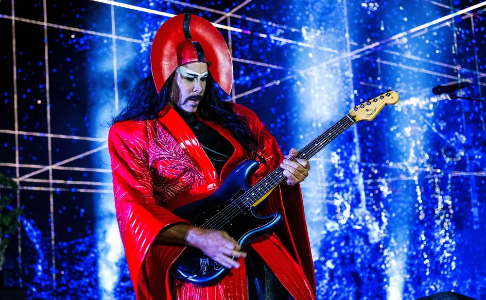 A person with striking makeup and a red headdress plays a black electric guitar on a stage with blue, sparkly lighting.