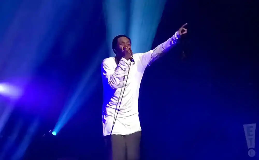 A man in a white shirt and gray pants sings into a microphone on a blue-lit stage, pointing his finger upwards.