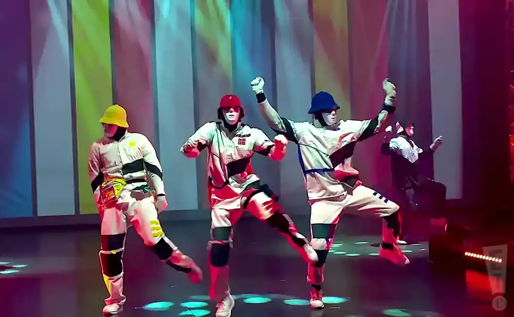 Dancers in colorful outfits and masks perform on a stage with a striped backdrop.