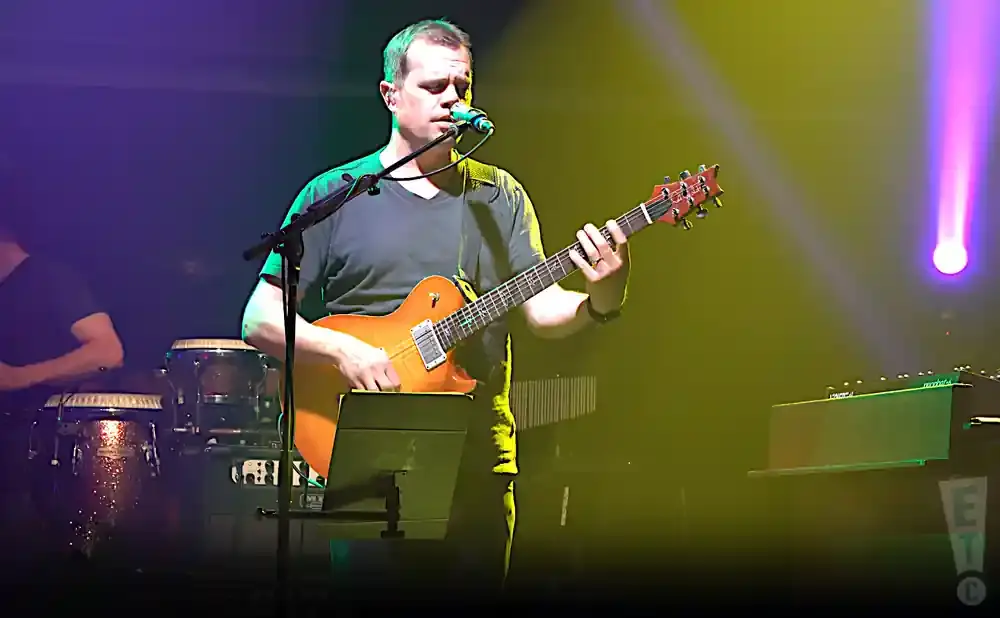 A man plays an orange electric guitar and sings into a microphone on stage with colorful lights.