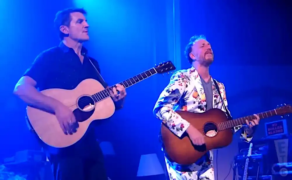 Two men playing acoustic guitars on a stage with blue lighting.
