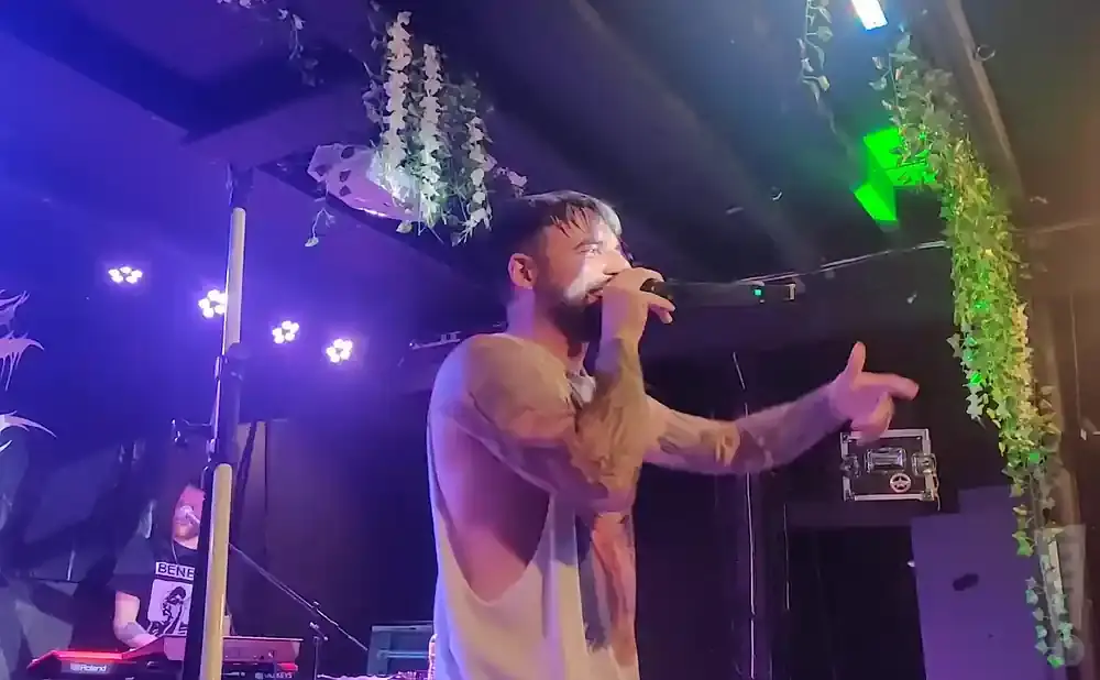 A tattooed man sings into a microphone on a stage lit by purple and green lights, with hanging ivy decorations.