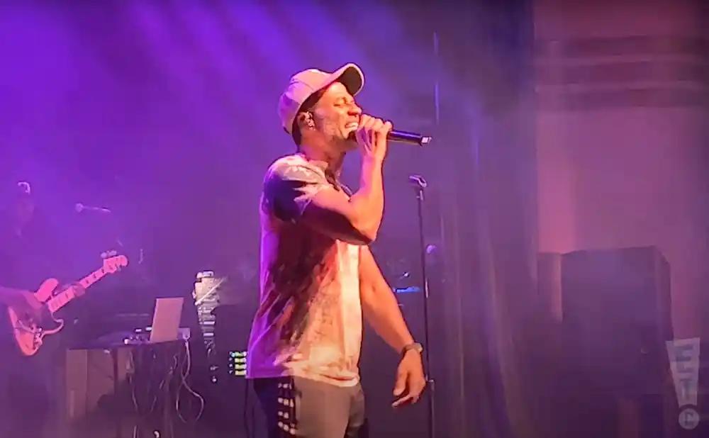 A man in a baseball cap and t-shirt sings into a microphone on a stage with purple lighting.