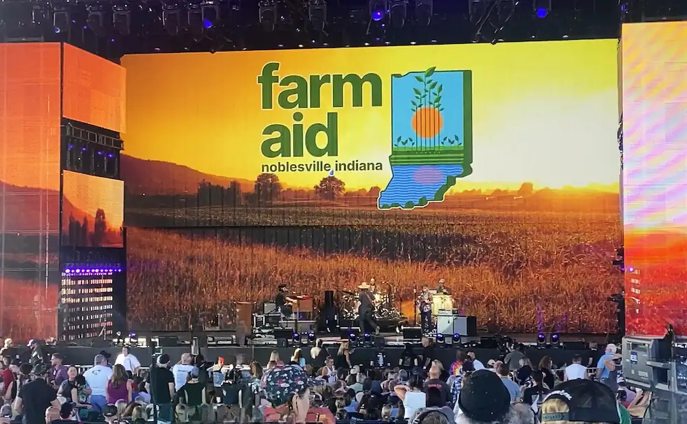 Farm Aid concert in Noblesville, Indiana, with a large screen displaying the Farm Aid logo and a scenic field at sunset.