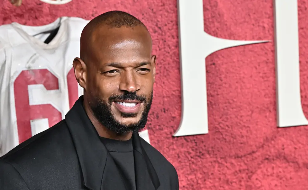 Maricon Wayans smiles in a black suit, with a white jersey bearing the number 6 blurred behind him on a red textured background.