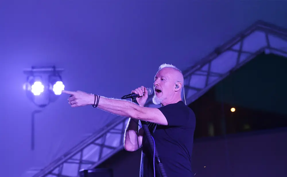 Man with white hair singing and pointing, illuminated by blue stage lights.