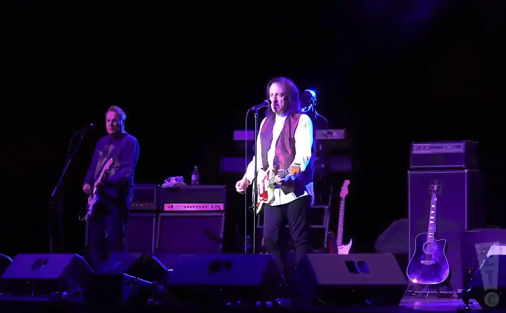 Two guitarists perform on a dark stage under purple lights. The guitarist on the left wears a blue shirt and plays a white guitar.