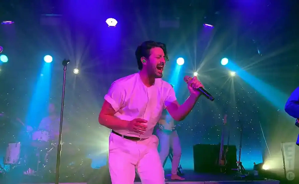 A male singer in white performs on stage, bathed in blue and purple lights.