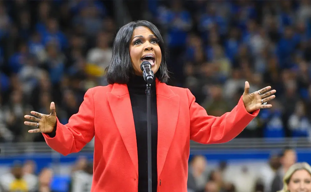 Singer CeCe Winans performs in a red blazer and black turtleneck, with hands extended.