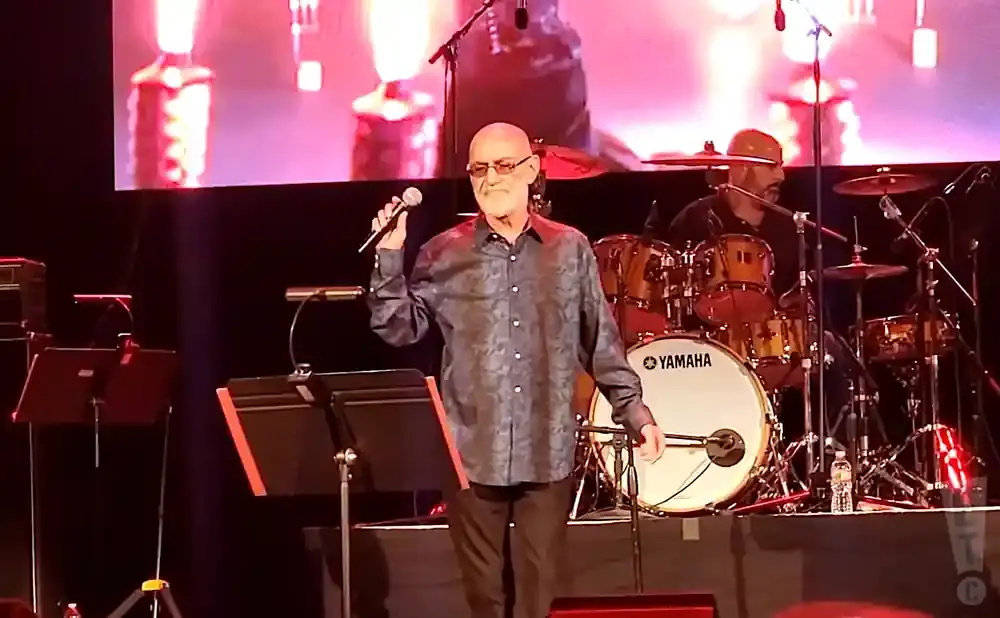 A man with glasses and a patterned shirt sings into a microphone on stage, with a drummer in the background.