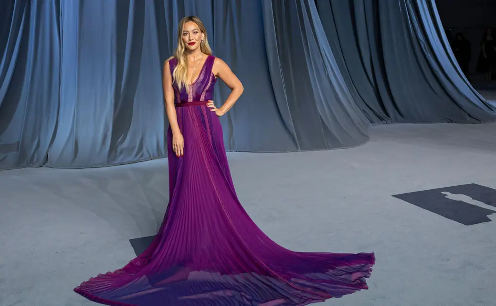 A woman in a flowing purple gown stands with her hand on her hip.