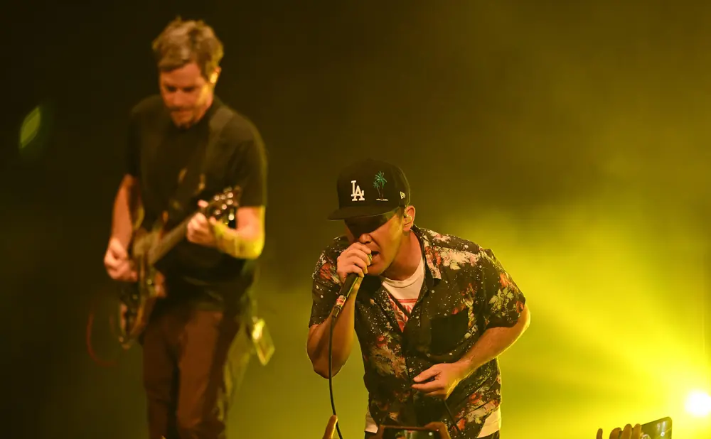 A musician with a LA Dodgers cap sings into a microphone on a stage lit by yellow lights.