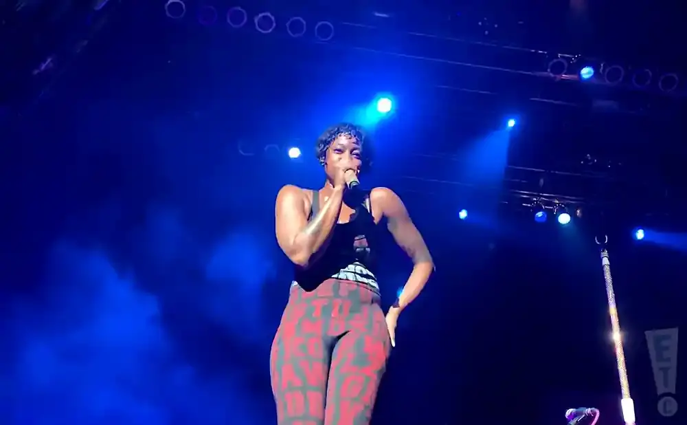 A woman in patterned leggings sings into a microphone on a dark stage lit by blue spotlights.