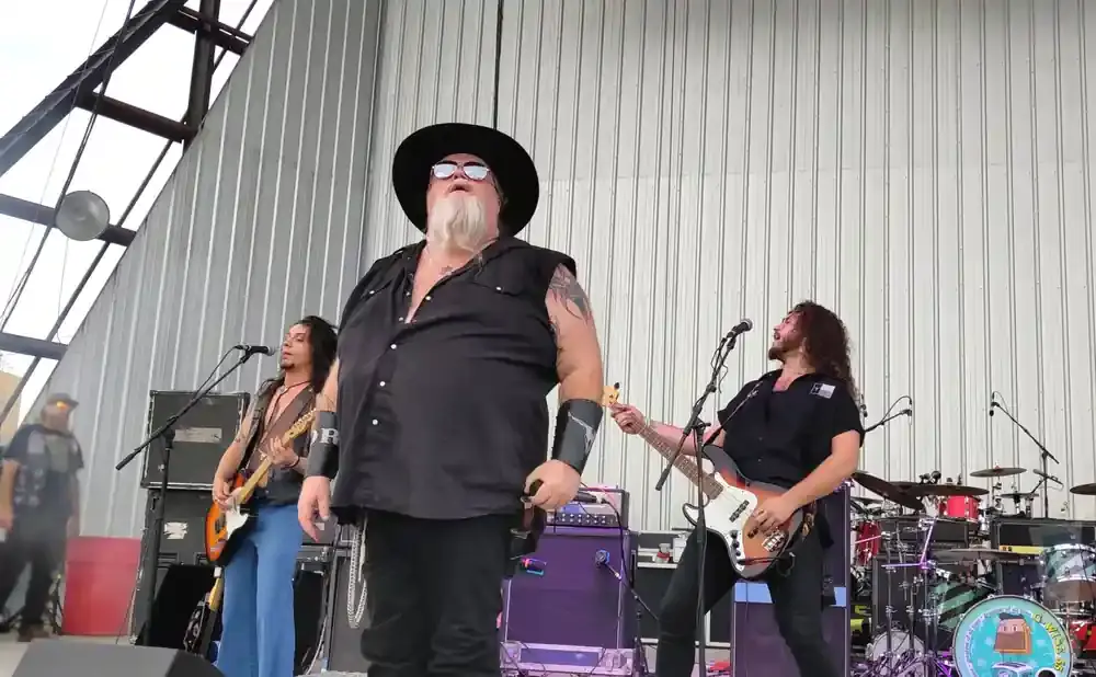 A band performs on an outdoor stage in front of a corrugated metal wall.