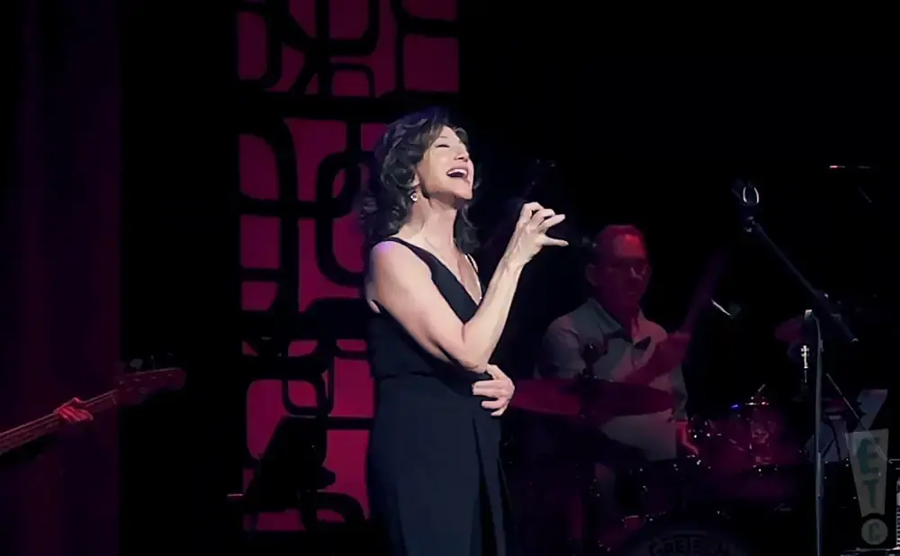 Singer Amy Grant performs in a black dress on stage, with a drummer and bassist visible.
