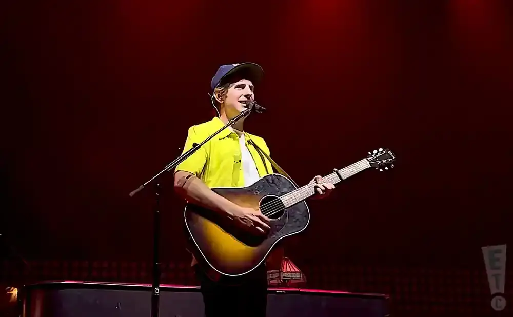 A man in a yellow shirt and blue baseball cap plays an acoustic guitar on stage.