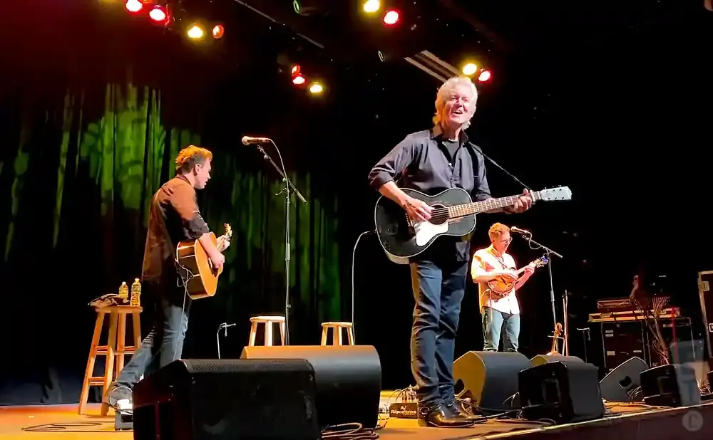 A band performs on stage; three musicians are visible, two with guitars and one with a mandolin.