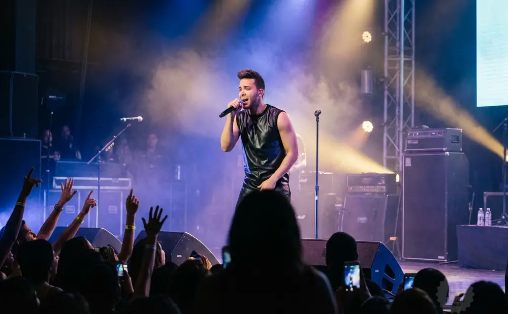 A male singer in a black leather outfit performs on stage, holding a microphone, with a crowd in the foreground.