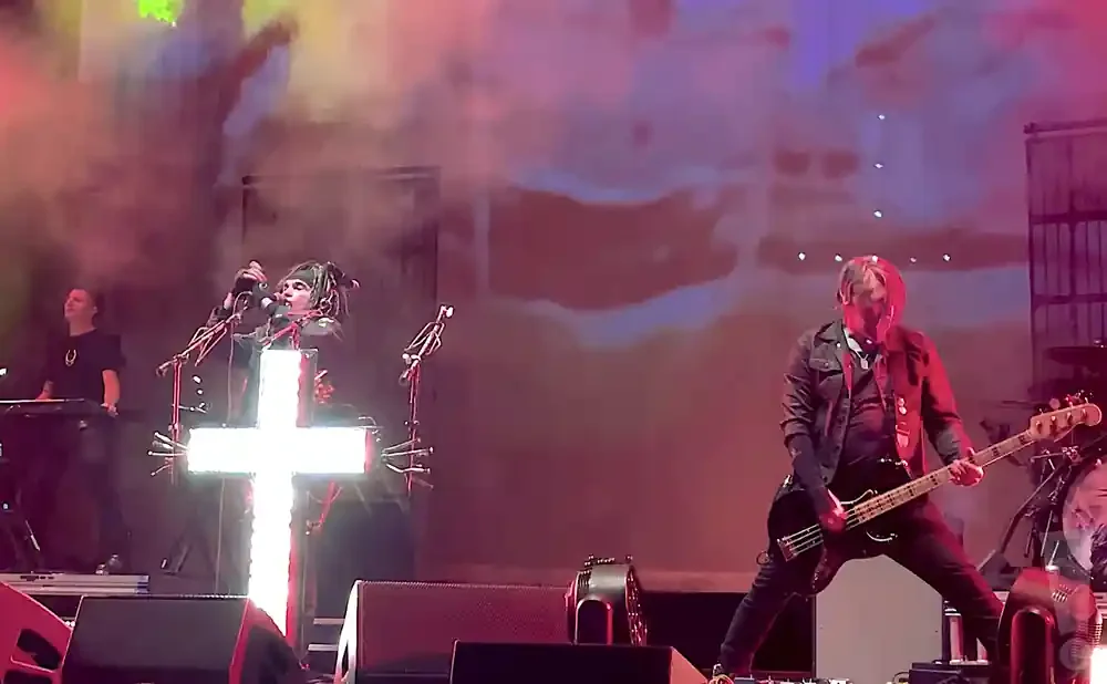 A band performs on stage with bright lights, a lit cross, and smoke effects.