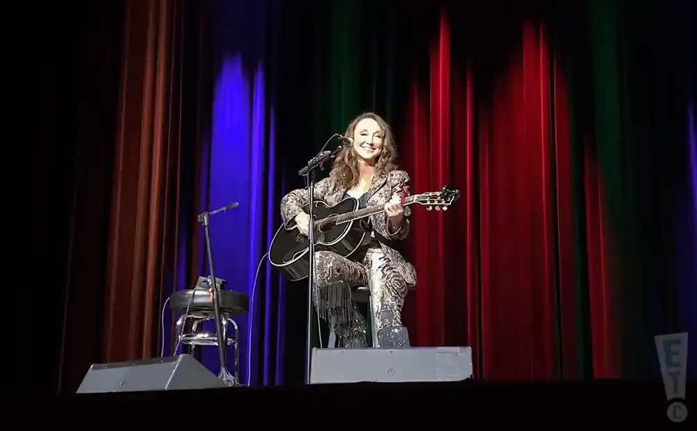 A woman in a sparkly outfit plays an acoustic guitar on stage in front of colorful curtains.
