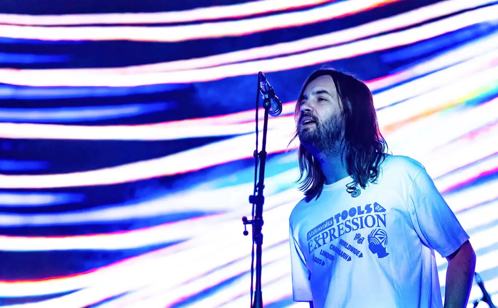 A man with long hair and a beard sings into a microphone on a stage with a colorful, striped background.