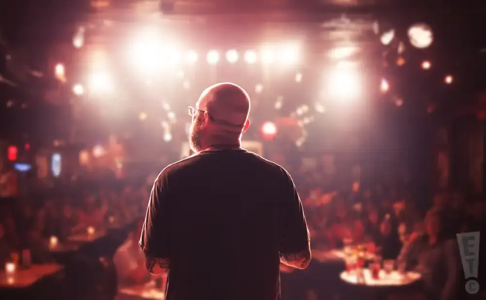 A bald man with glasses on a stage faces a blurred audience under bright stage lights.