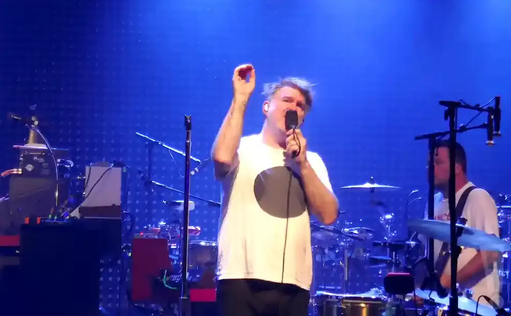 A man in a white t-shirt sings into a microphone on a stage with a blue backdrop.
