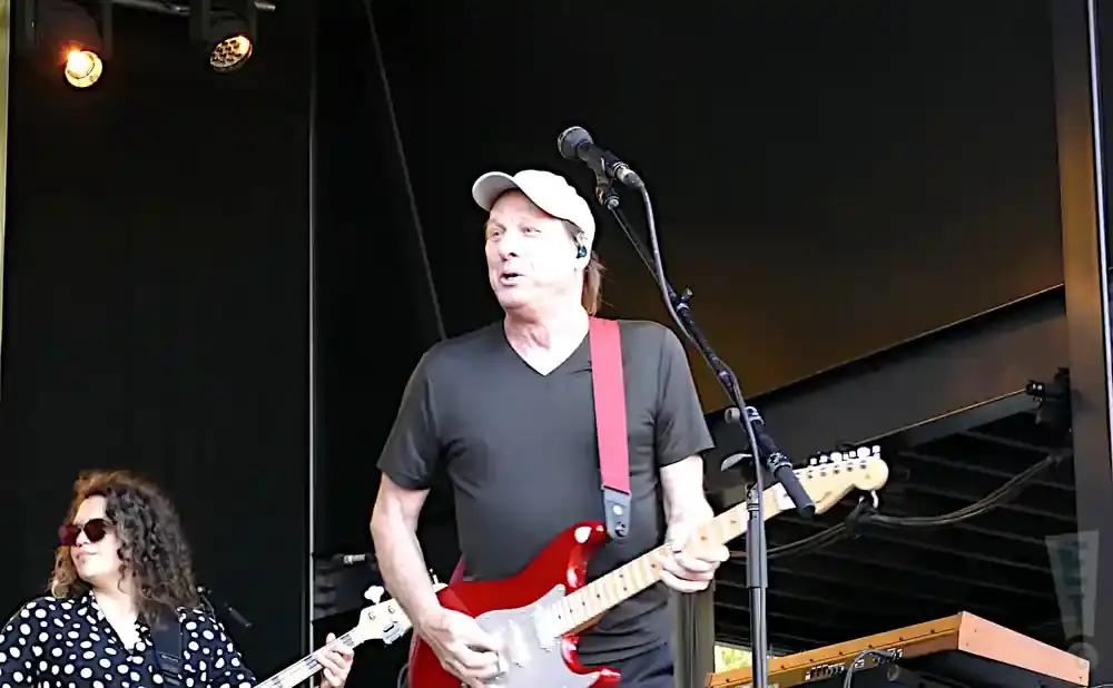 A man in a white cap and dark shirt plays a red electric guitar on stage.