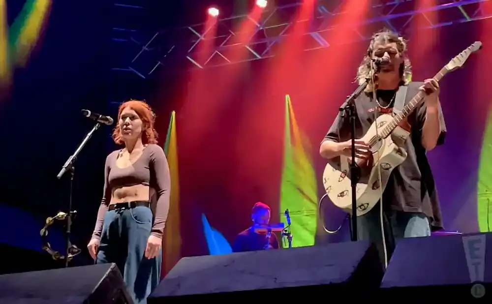 A woman with red hair sings into a microphone while a man plays guitar on a stage with colorful lighting.