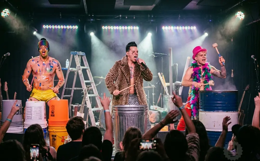 Three musicians perform on stage with colorful outfits and body paint, playing drums made of buckets and barrels.