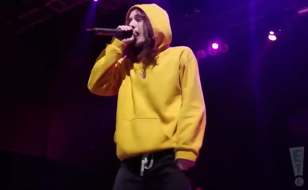 A person in a yellow hoodie raps into a microphone on a dimly lit stage with purple lighting.