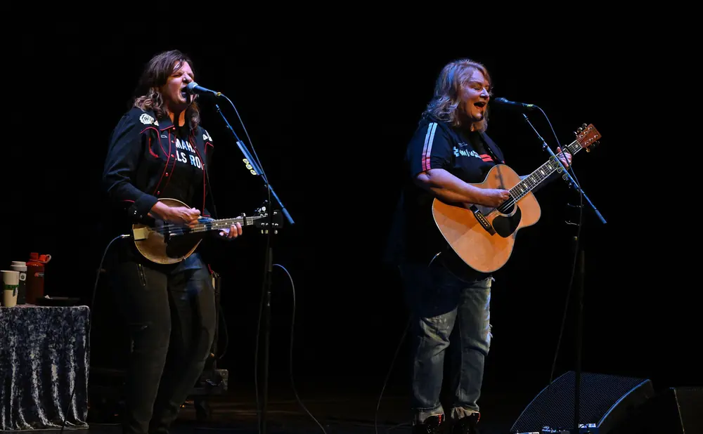 Two women playing instruments and singing into microphones on a dark stage.