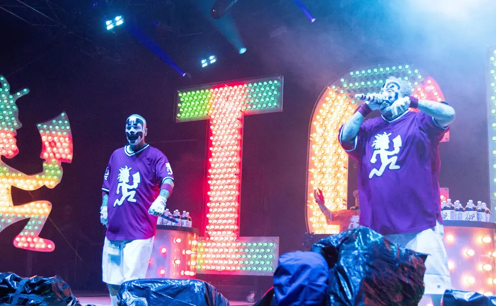 Performers in clown makeup and purple jerseys on a stage with lit signs and colorful lights.