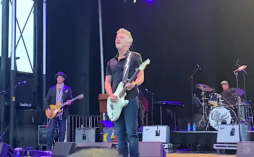 A rock band performs on an outdoor stage with a black backdrop. A guitarist in the foreground plays a white electric guitar.