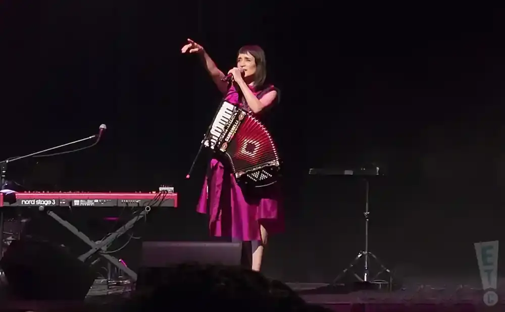 A woman in a pink dress plays an accordion on stage next to a keyboard.