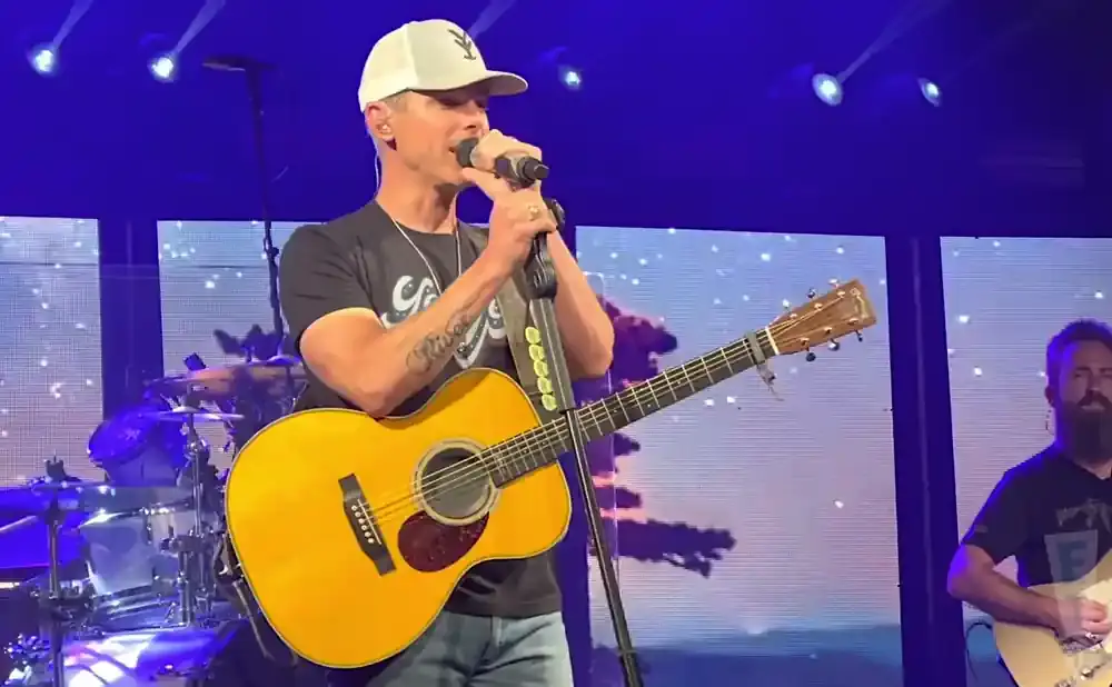 A male musician in a white cap and black t-shirt sings and plays a yellow acoustic guitar on stage.