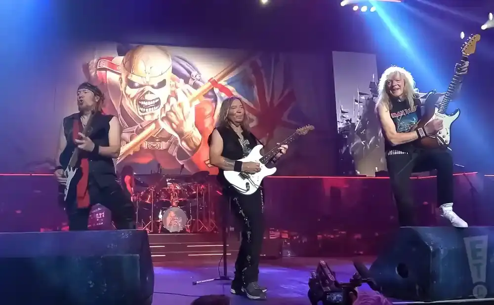 Iron Maiden performs on stage in front of a large backdrop featuring the band's mascot, Eddie, and the Union Jack flag.