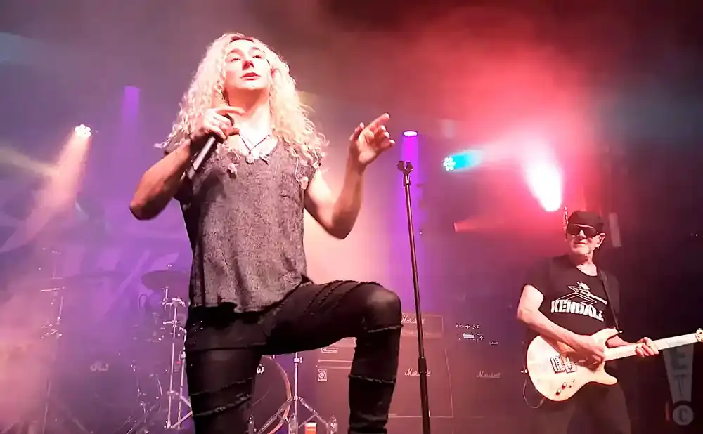 A male singer with long blonde hair performs on stage with a guitarist in the background.