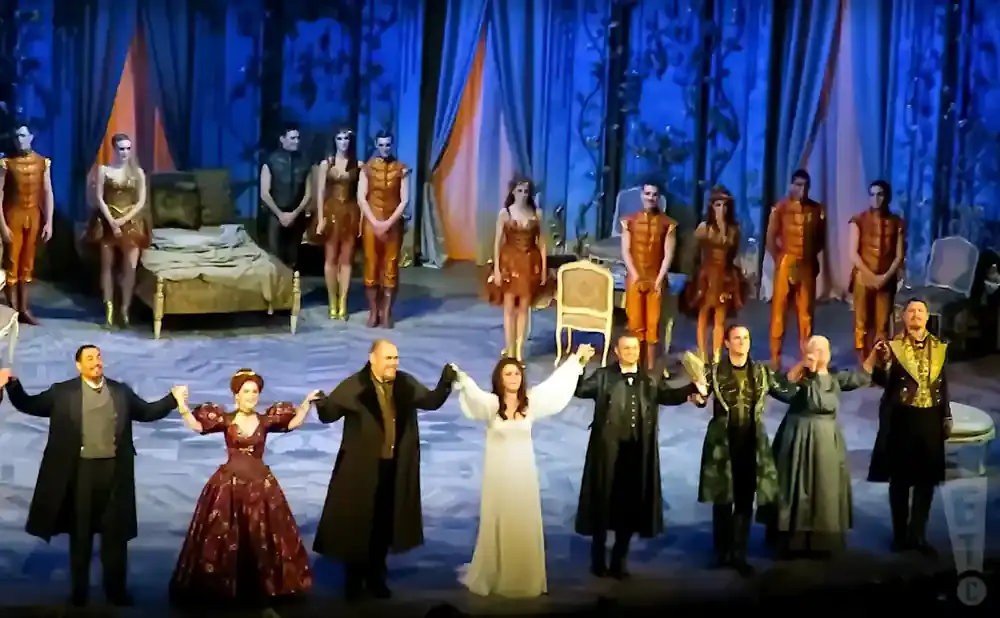 A theater cast takes a bow on stage, with actors in ornate costumes and a bed on the set.