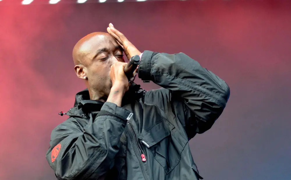 Freddie Gibbs performs on stage, wearing a black jacket and holding a microphone to his mouth with his hand on his forehead.
