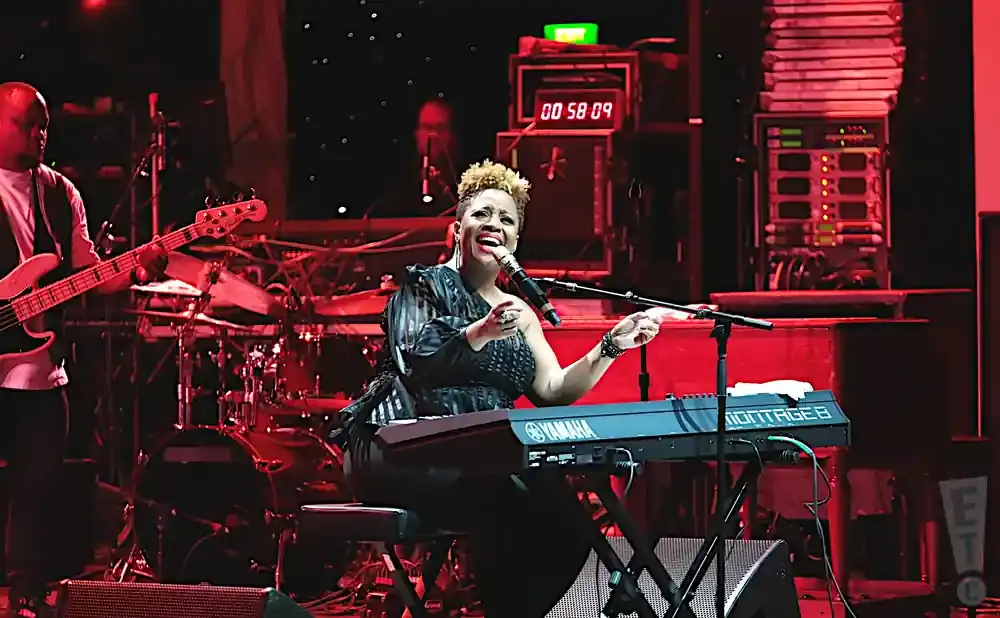 A female keyboardist sings into a microphone while playing a Yamaha keyboard on stage with a band.