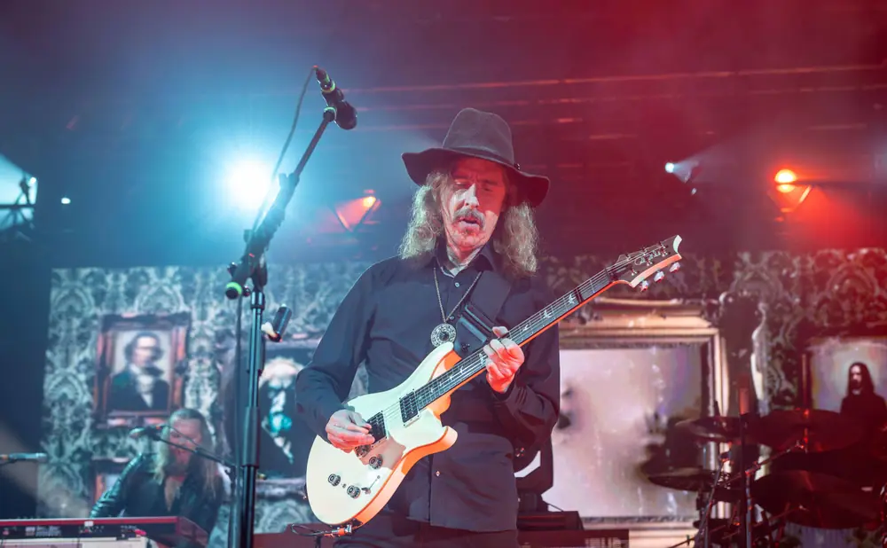 A male guitarist in a black hat and shirt plays a white electric guitar on stage with red and blue lighting.
