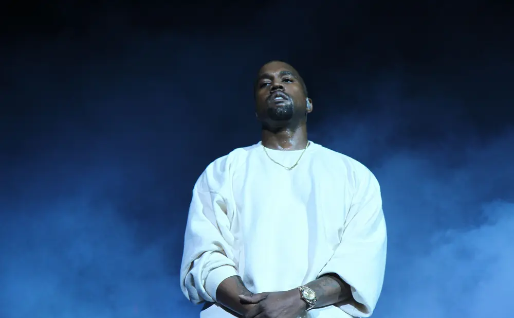 Kanye West stands on stage wearing a white sweatshirt and a gold chain.