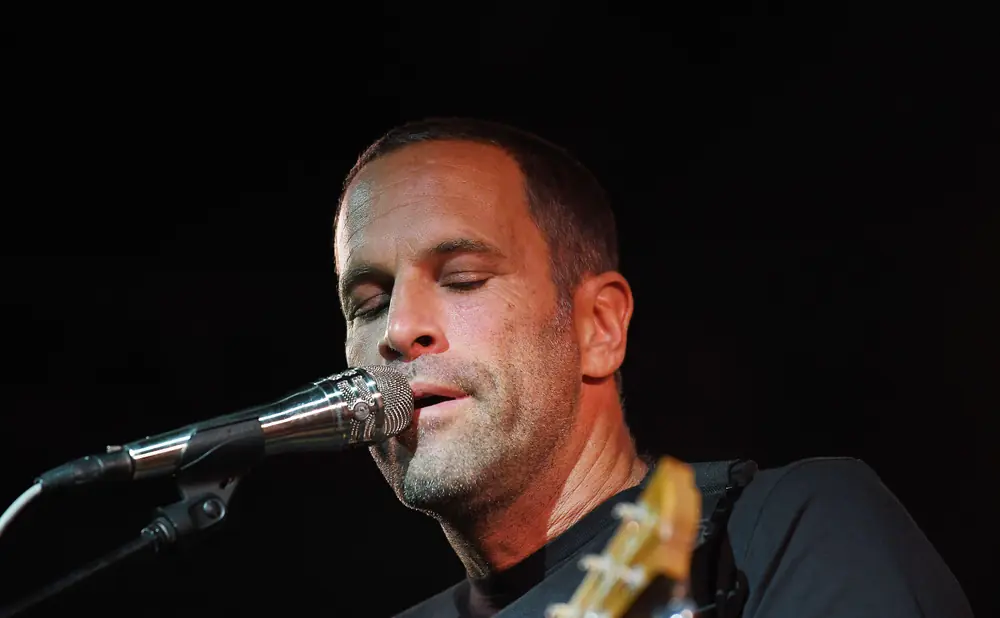 A man plays an acoustic guitar and sings into a microphone on a stage with blue lighting.