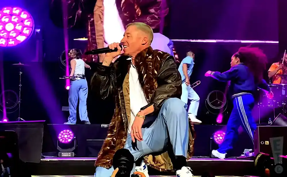 A male singer on stage wearing a long patterned jacket and blue pants, holding a microphone.