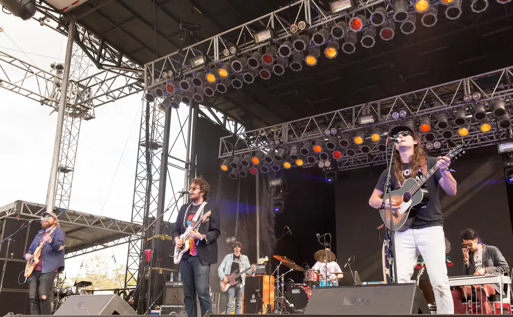 A band plays on an outdoor stage with a large lighting rig overhead and many speakers on the ground.