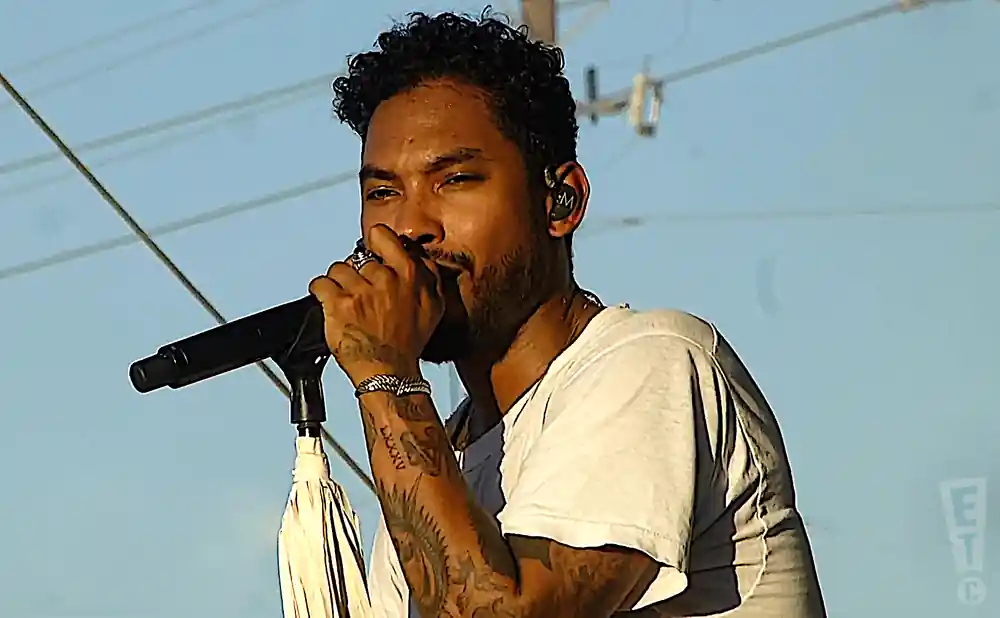 Singer Miguel performing on stage, holding a microphone and wearing a white t-shirt.
